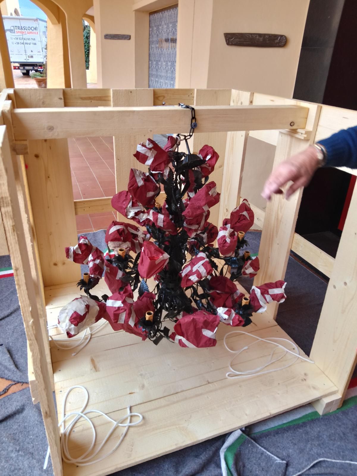Una composizione floreale rossa è sospesa in una cassa di legno, con la mano di una persona che la indica.