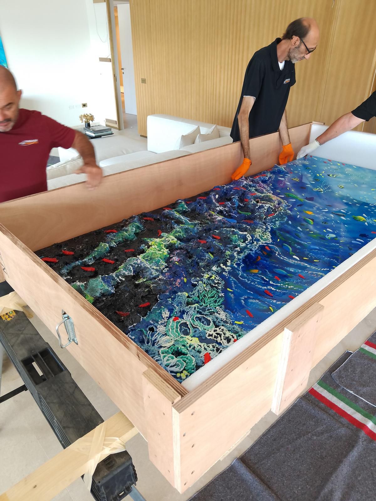 Tre uomini estraggono con cautela un grande dipinto colorato a tema oceanico da una cassa di legno.