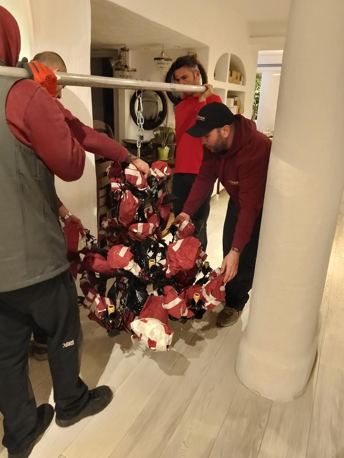 Tre persone con magliette rosse spostano un grande oggetto decorato con colori rossi e bianchi all'interno di un edificio.