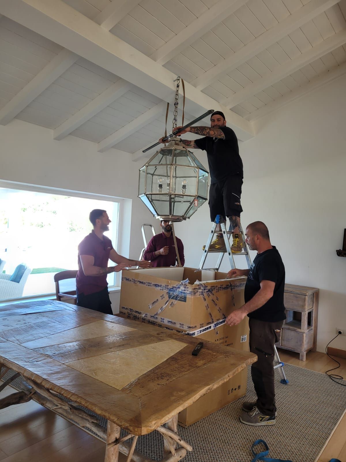 Tre uomini installano una grande lampada sopra un tavolo da pranzo; due su una scala, uno a terra. Stanza luminosa.