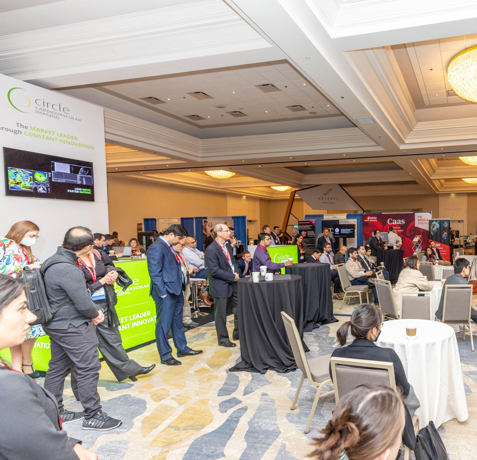 Conference room with attendees, speakers, and booths. Green and white signage, people conversing, round tables.