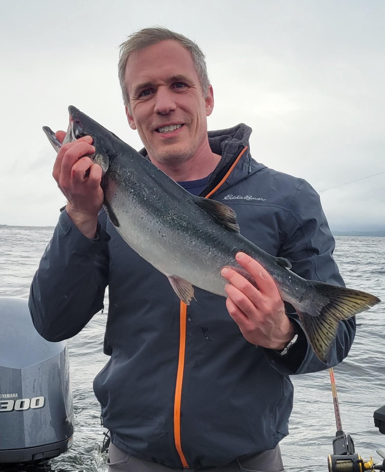 Man smiling, holding a silver fish on a boat. Gray sky and water. Blue jacket with orange zipper.