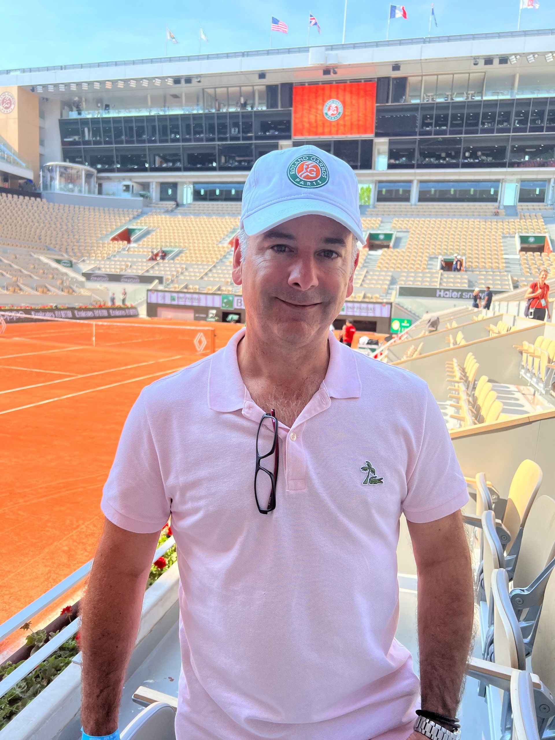 Man in a pink polo shirt and white hat at a tennis stadium with an orange clay court.