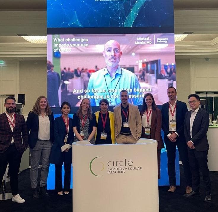 Group of people posing in front of a Circle Healthcare Marketing display; video screen in background.