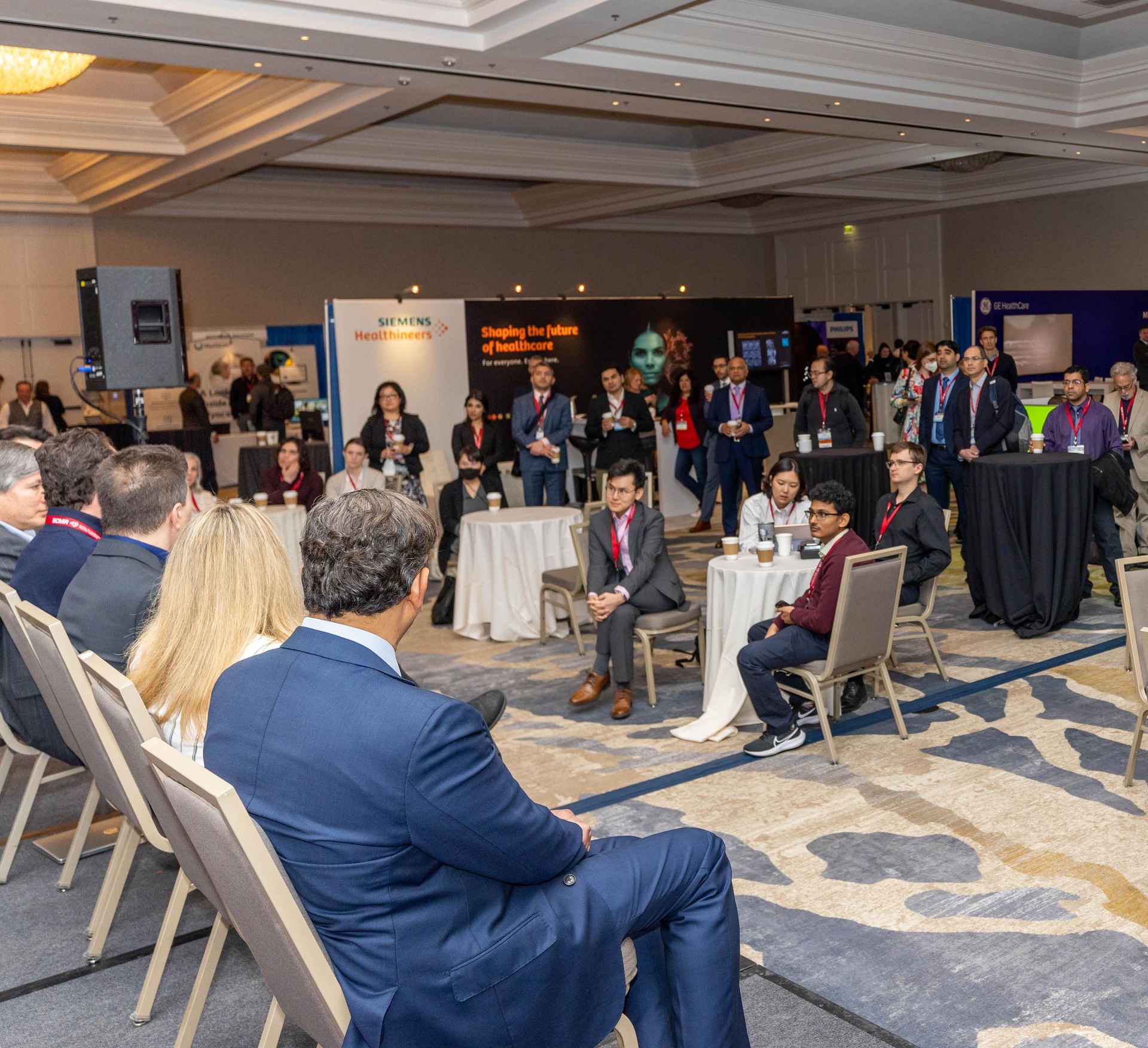 Conference attendees in a large room; some seated, others standing and talking.
