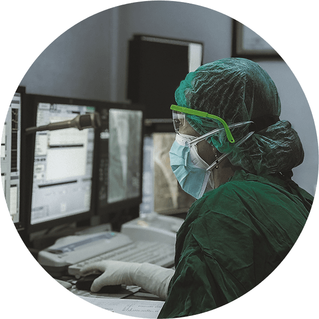 Medical professional in green scrubs, mask, and goggles operates a computer with medical scans displayed.