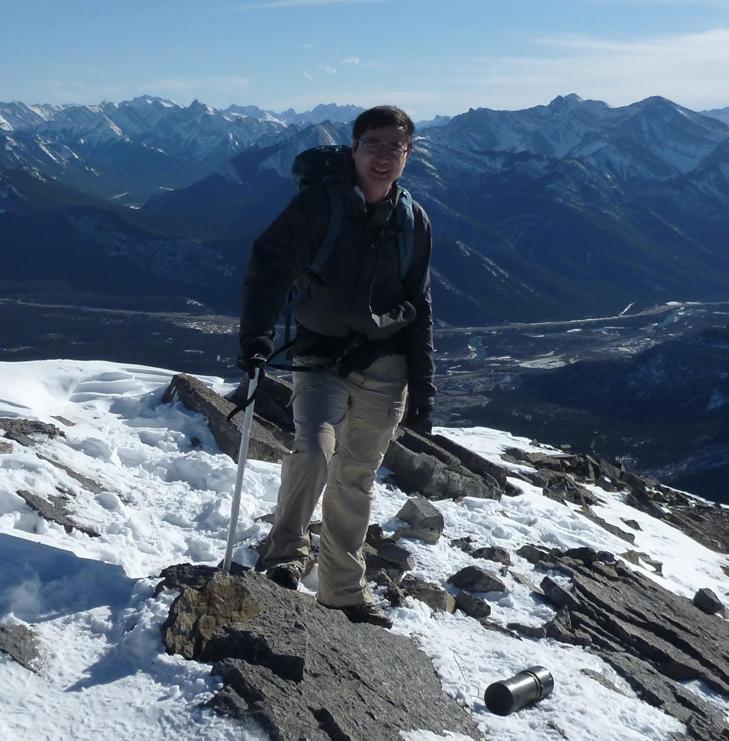 Person on a snowy mountain peak, wearing backpack, holding a hiking stick, overlooking valley and mountains under a blue sky.