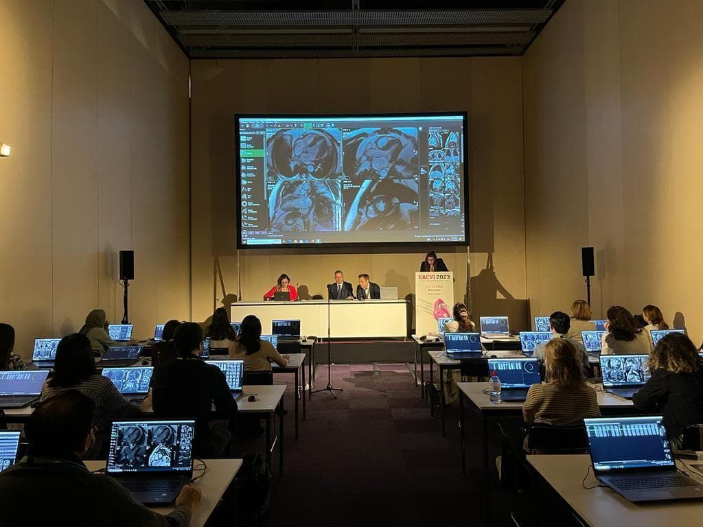 Conference room with attendees at laptops facing a screen displaying medical images; speakers at the front.