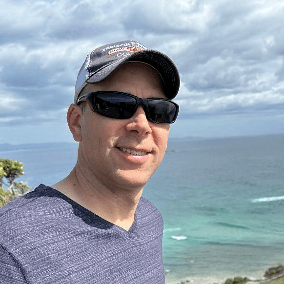 Man in sunglasses and hat smiles by the ocean, cloudy sky background.