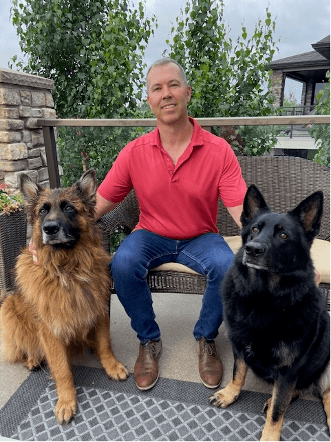 Man sitting with two German Shepherds on a patio. He wears a red shirt and jeans.