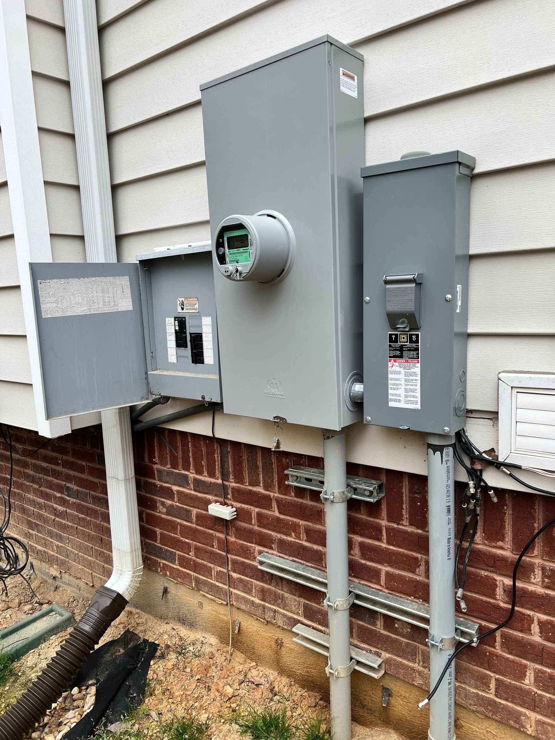 Exterior electrical panel box mounted on brick wall. Gray metal boxes, conduit, and meter socket. Open panel door.