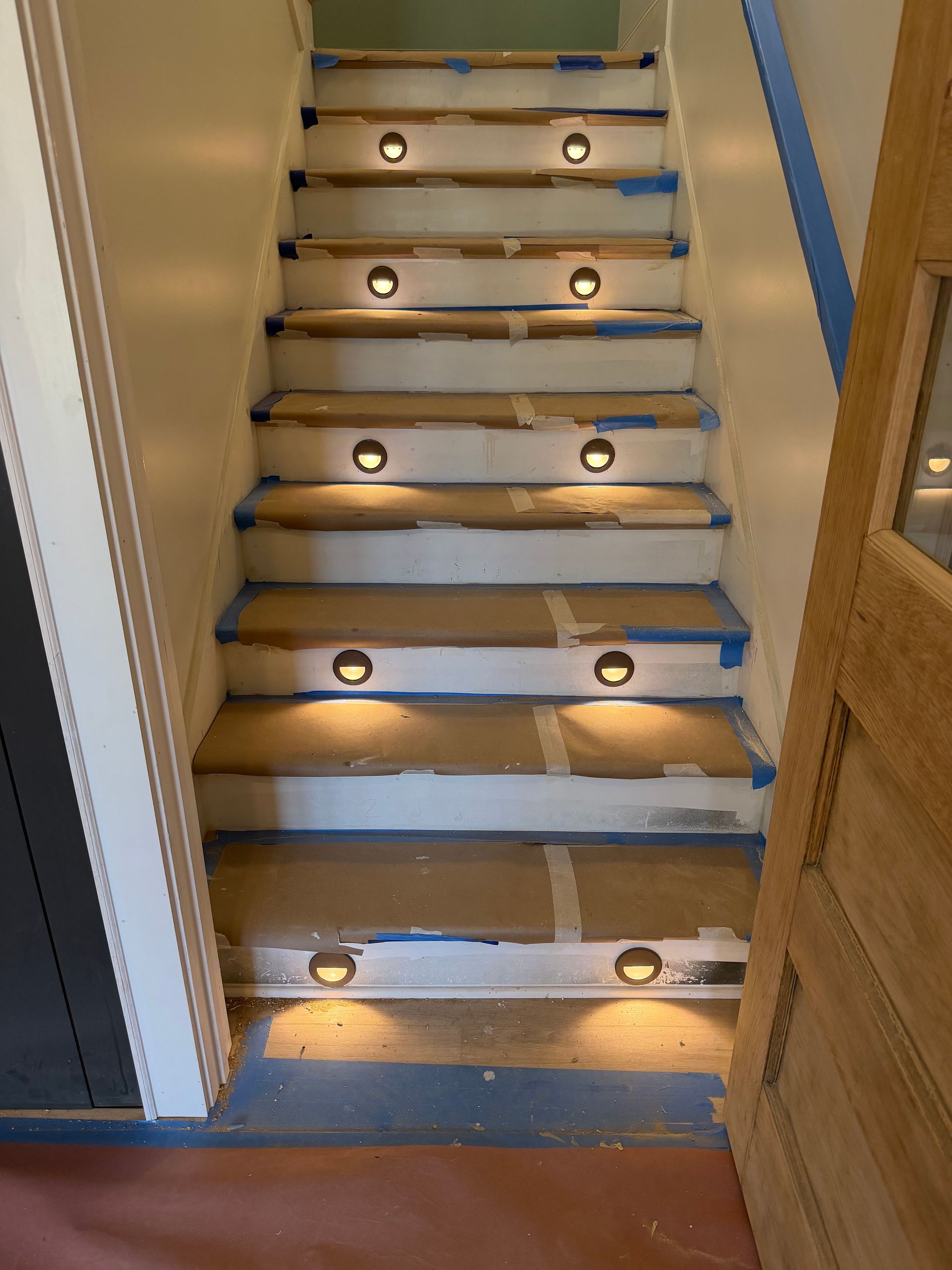 Staircase with recessed lights. Beige steps with blue painter's tape, lit by circular, warm lights. Door visible on the right.