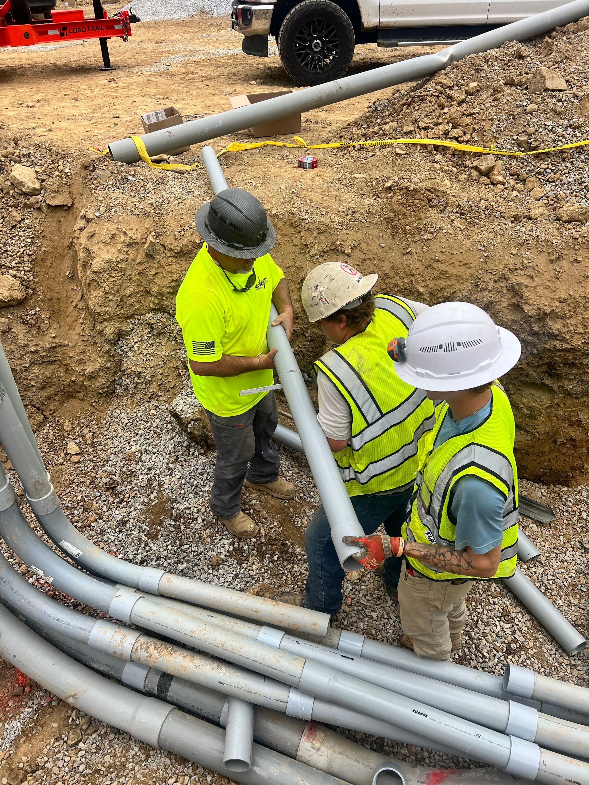 Three workers in hard hats and neon vests installing gray pipes in an open trench.