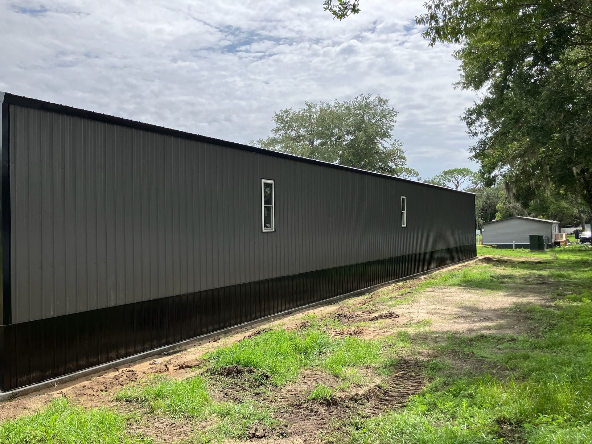 Dark gray metal-sided building with two windows, set on a grassy lot.
