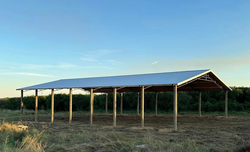 A large, open-sided wooden shelter with a metal roof stands in a field under a blue sky.
