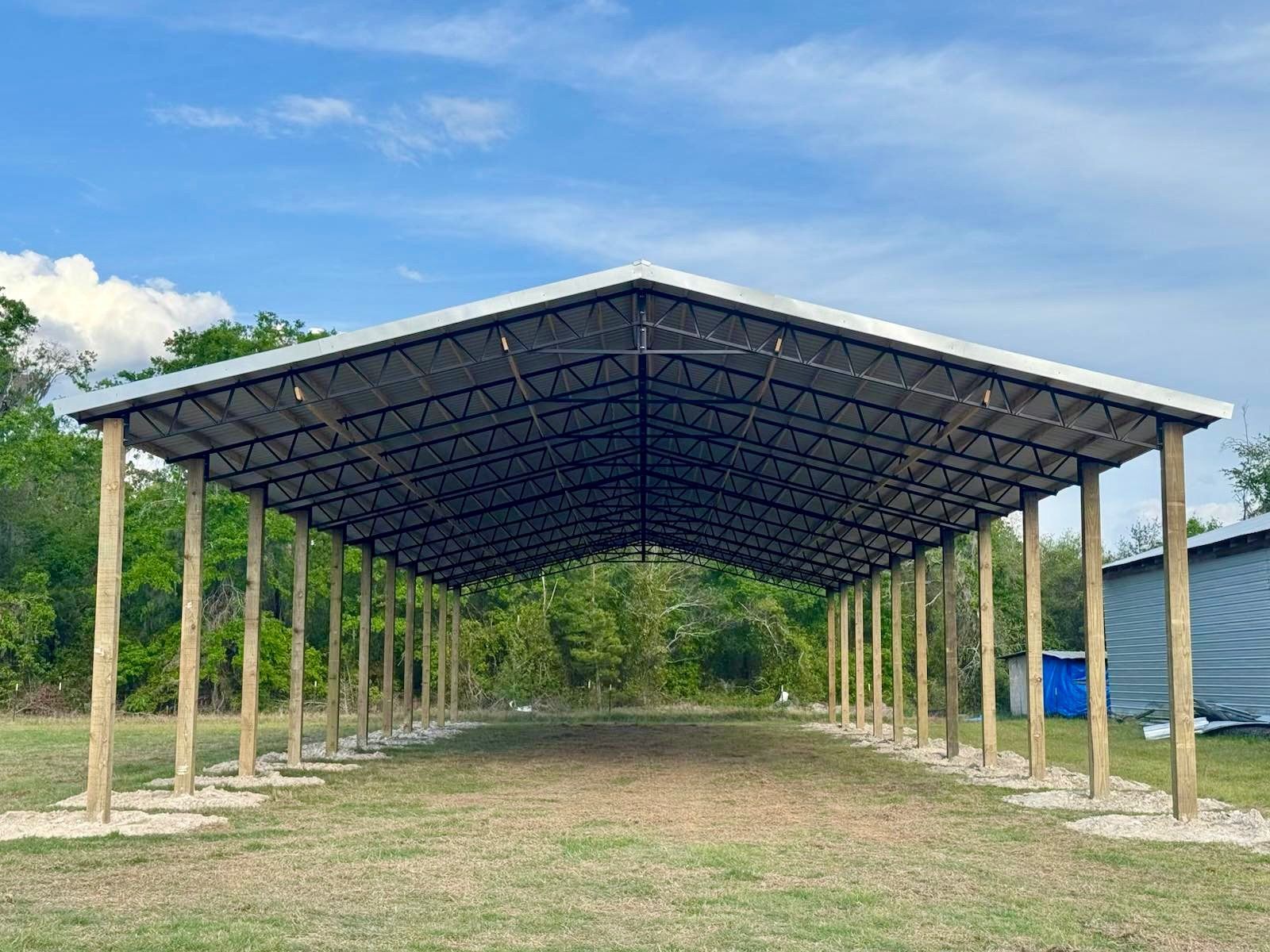 Large open-air shelter with a metal roof supported by wooden posts set in concrete, on a grassy field under a blue sky.