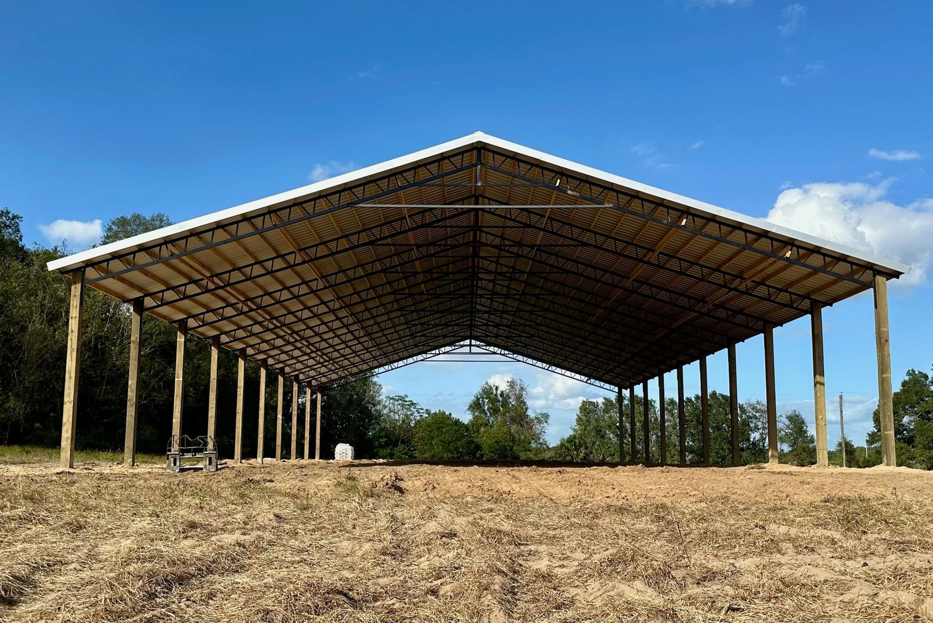 A large, open-sided metal and wood pavilion under a blue sky, with wood posts and a dirt floor.