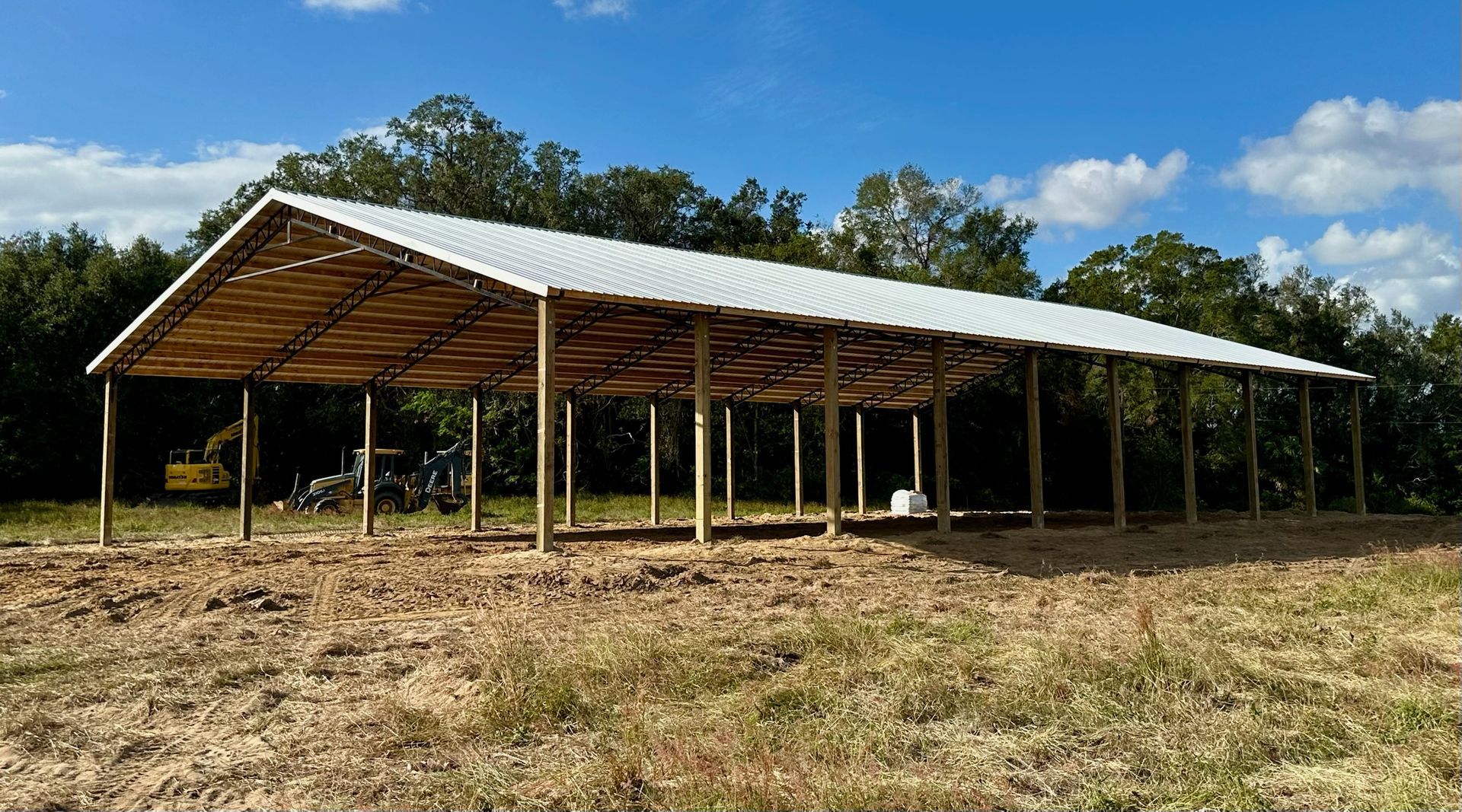 Large open-sided wooden structure with a white metal roof in a field. Blue sky and trees in the background.