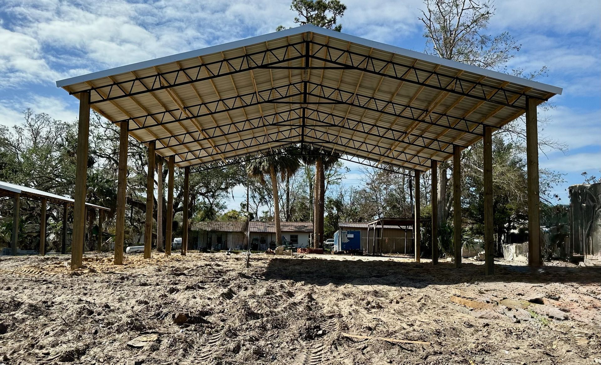 A large open-air metal shed with a dirt floor, supported by wooden posts, in a rural setting.