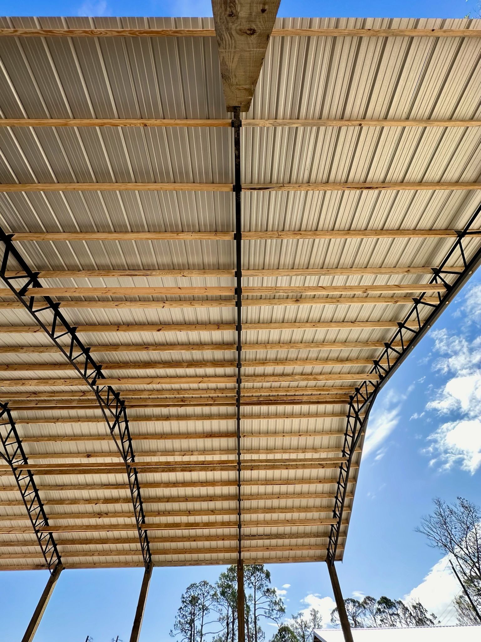 Looking upwards under a corrugated metal roof supported by wooden beams and dark metal truss work against a blue sky.