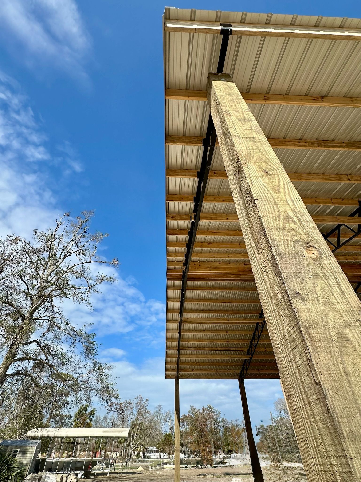 Wooden beam supporting a metal roof on a sunny day. Sky is visible with some tree branches.