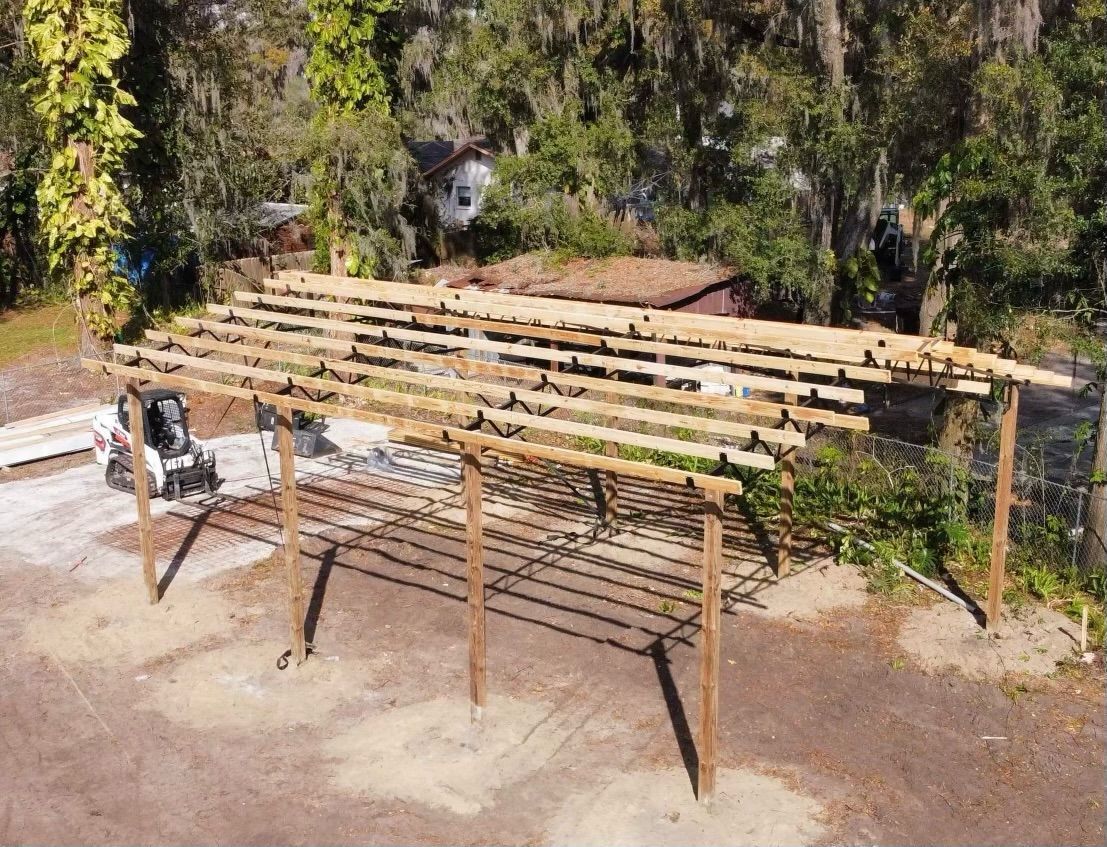 Wooden carport under construction; beams and supports visible, set in a yard.