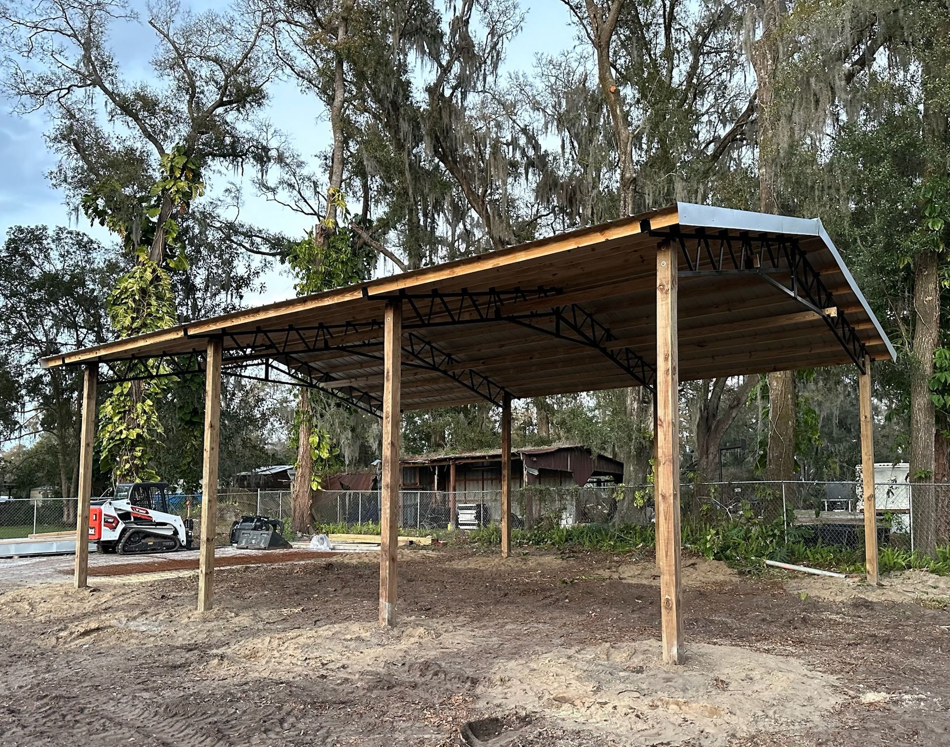 Open-air metal roof structure supported by wooden posts in a cleared area with trees in the background.
