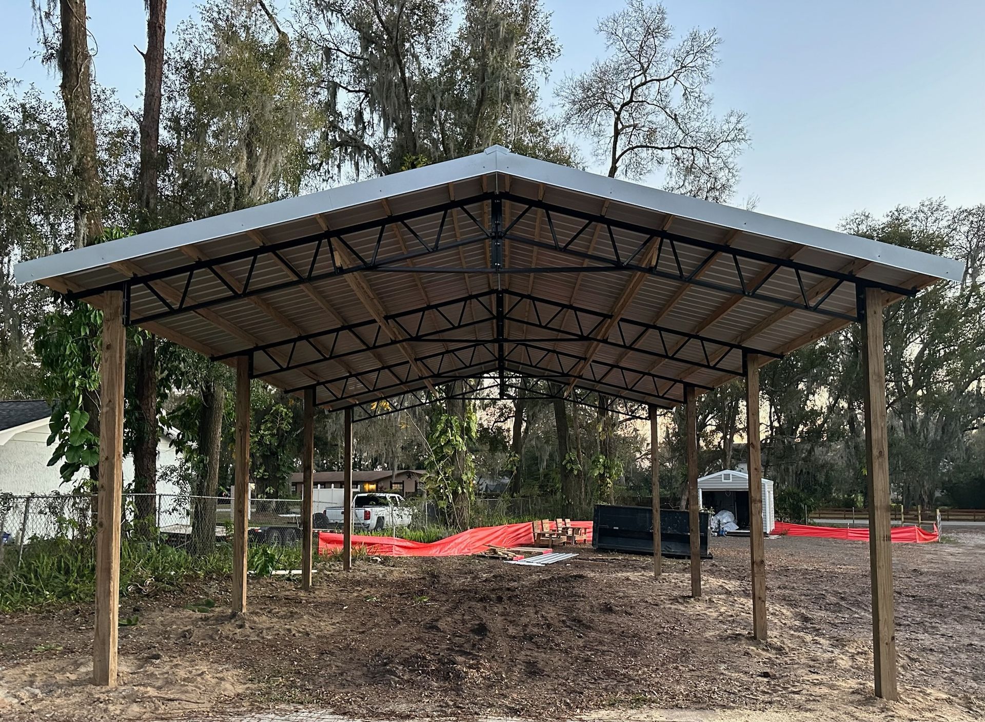 Open-air metal roof carport supported by wooden posts, situated outdoors on a dirt surface.