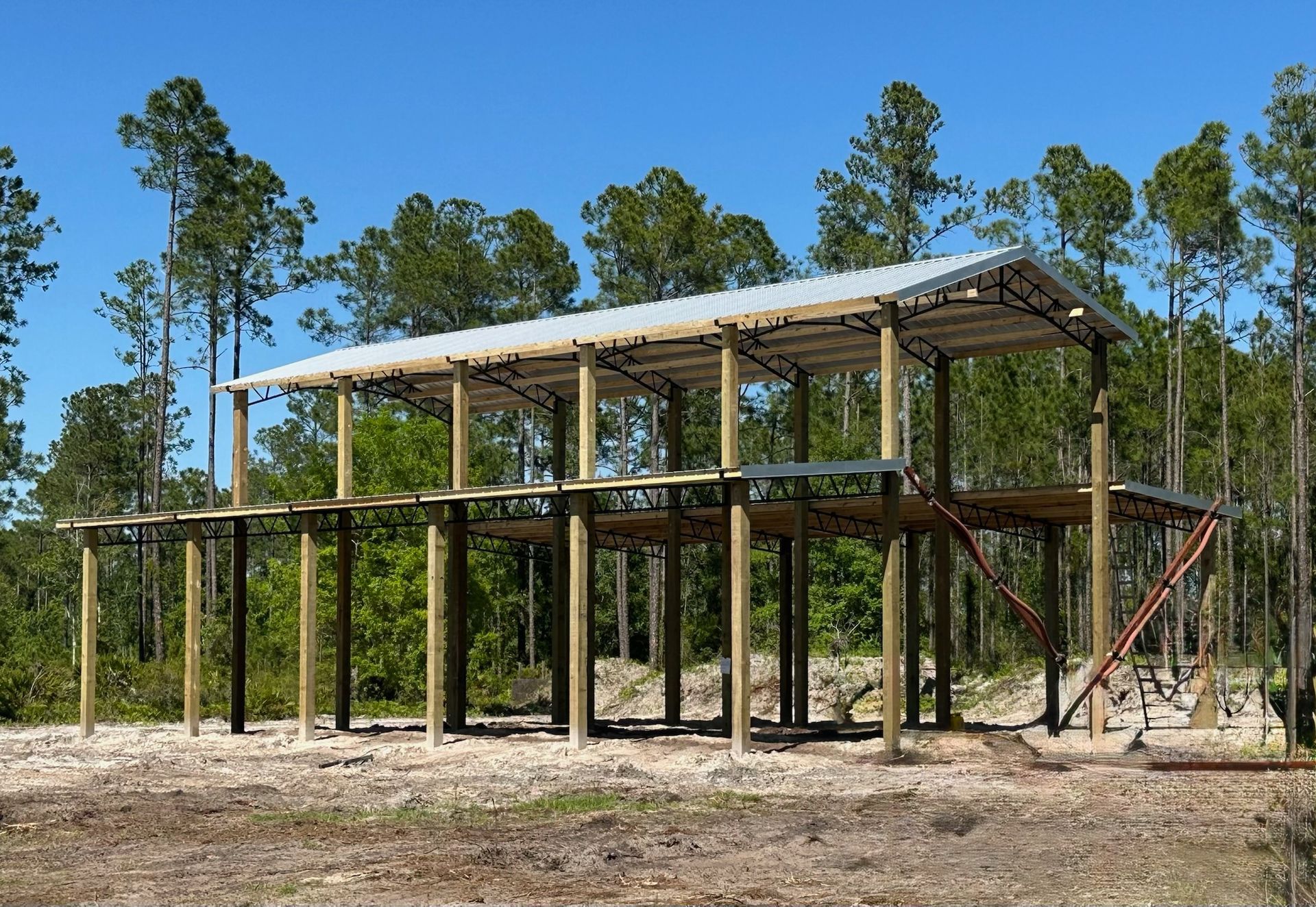 Two-story wooden structure under construction, with a metal roof, set against a forest and blue sky.
