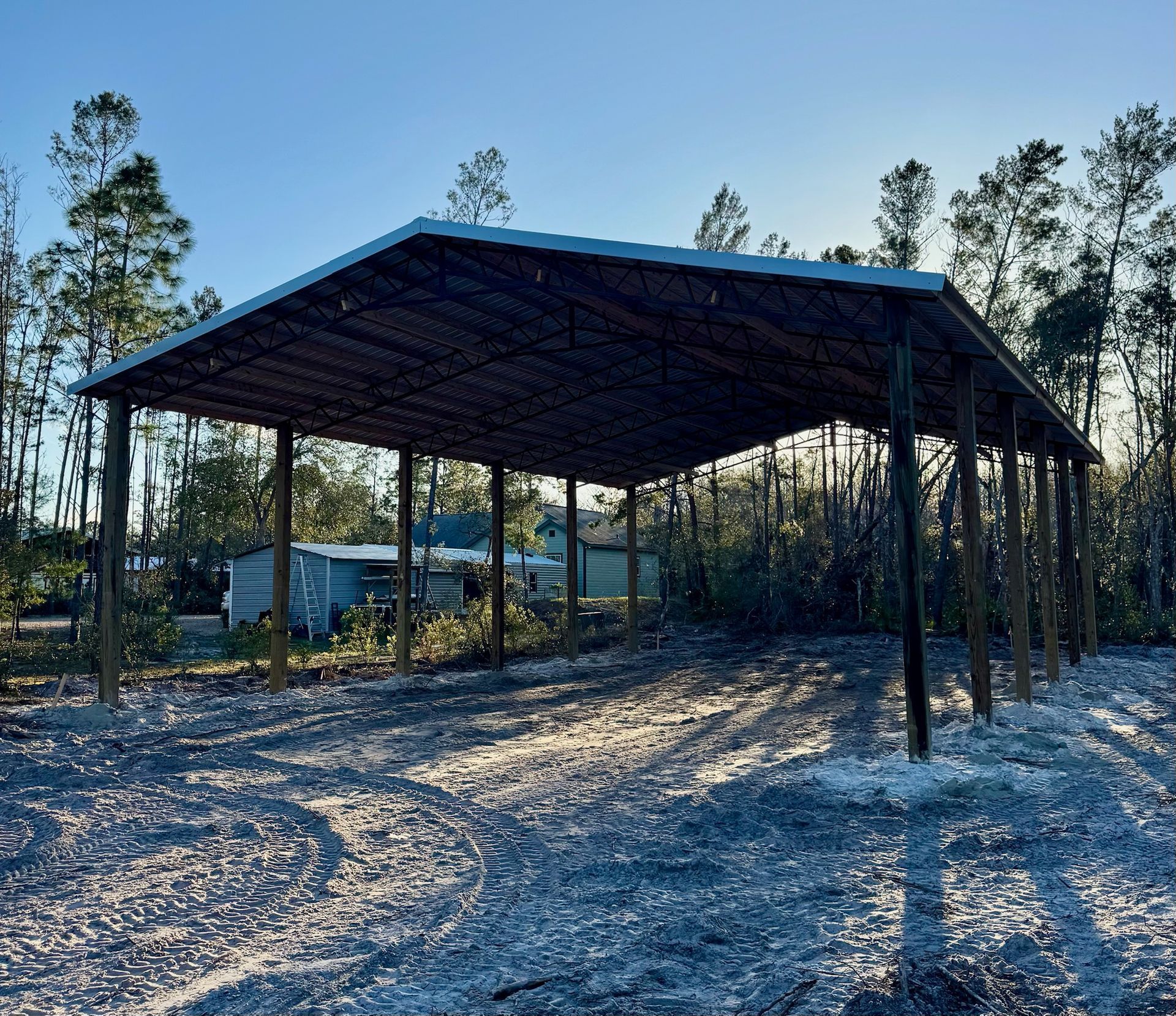Open-air metal carport with wooden posts, in a dirt clearing with trees, under a blue sky.