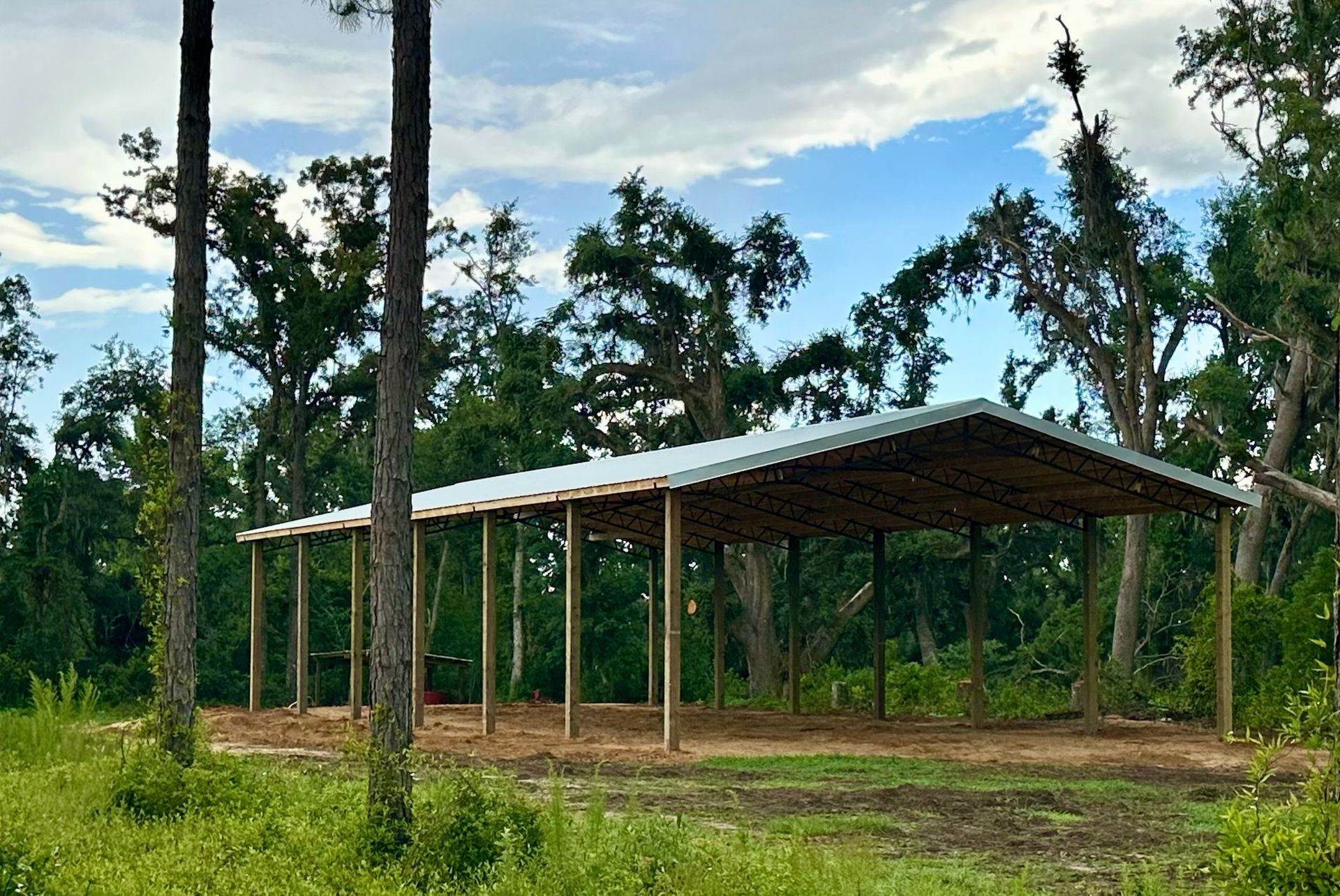 Carport with metal roof supported by wooden posts in a wooded area under a cloudy sky.