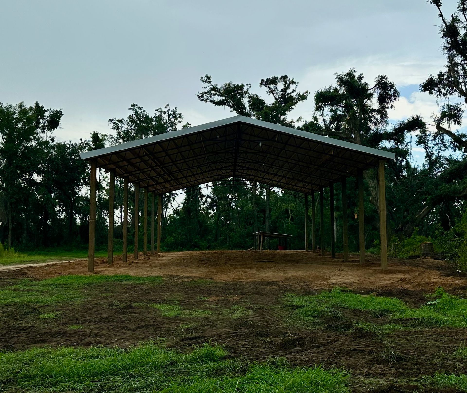 Open-air shelter with a metal roof, built on dirt. Wood posts support the roof. Green grass and trees are in the background.