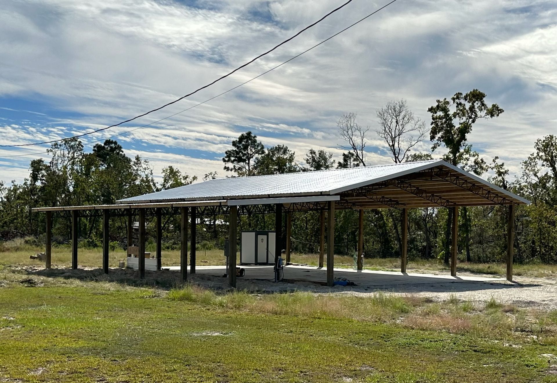 A wooden shelter with a metal roof in a grassy area, trees in the background, power lines overhead.