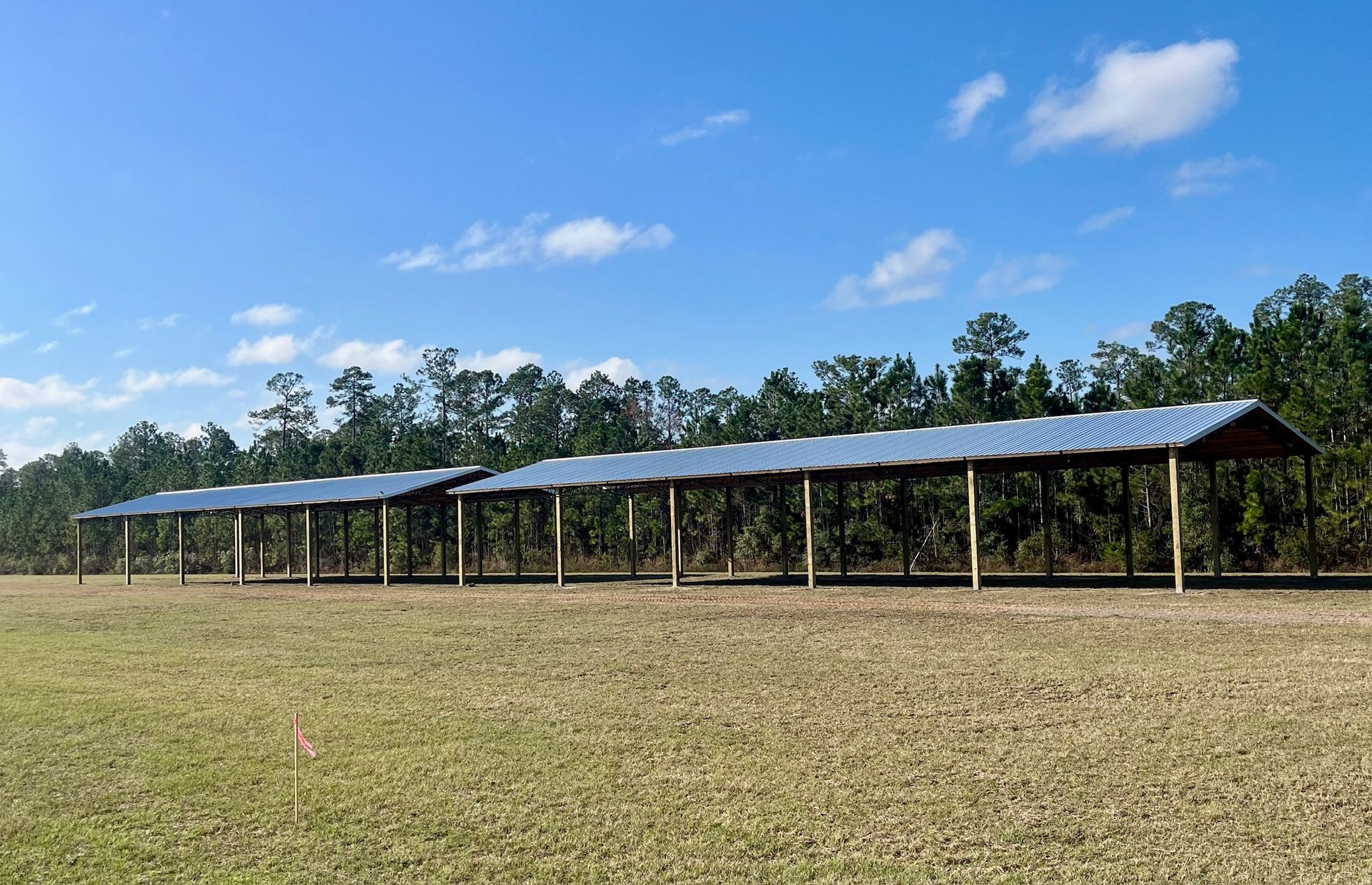 Two long, open-sided structures with metal roofs in a grassy field, trees in the background, blue sky.