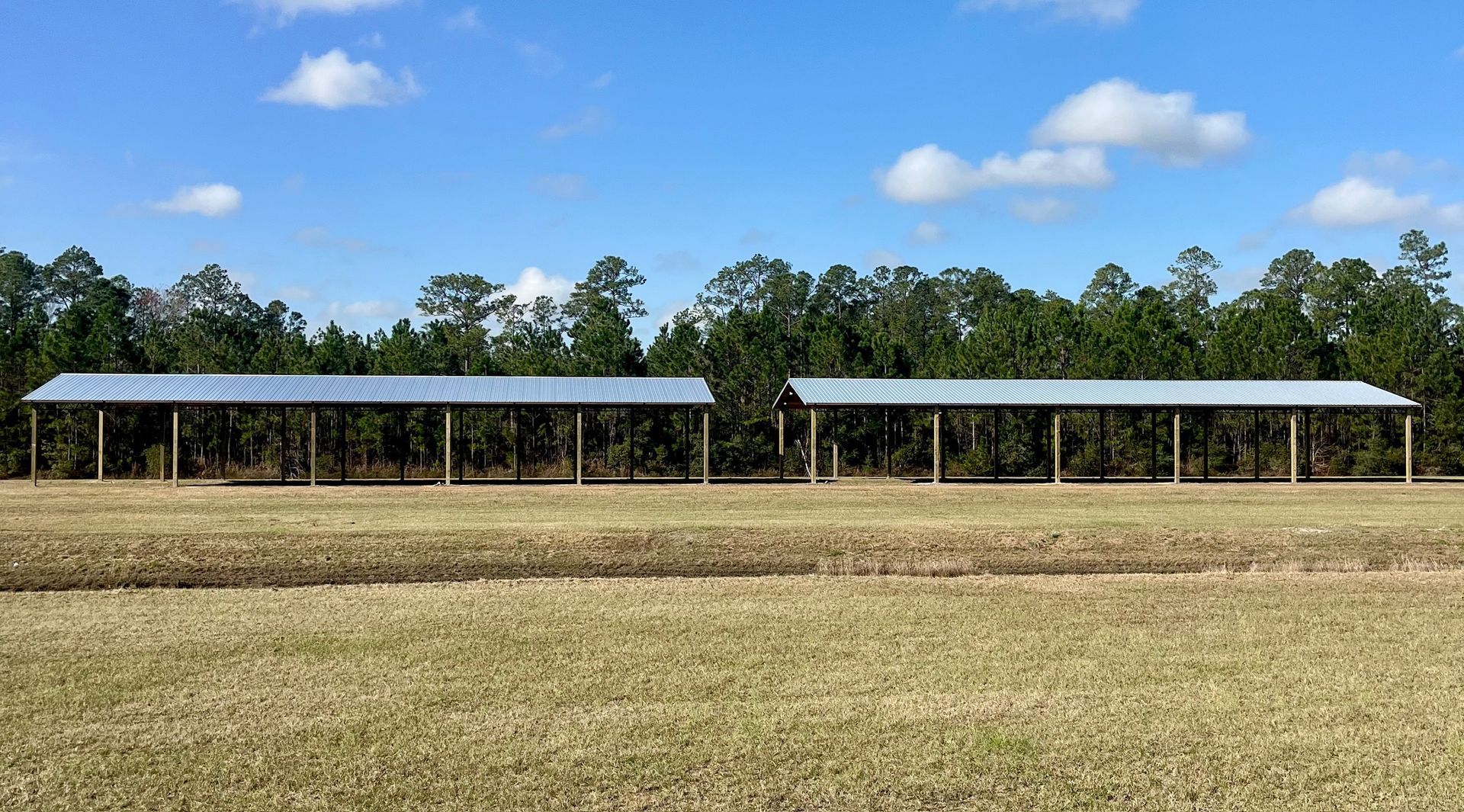 Two long, open-sided shelters with metal roofs in a field, trees in the background, blue sky.
