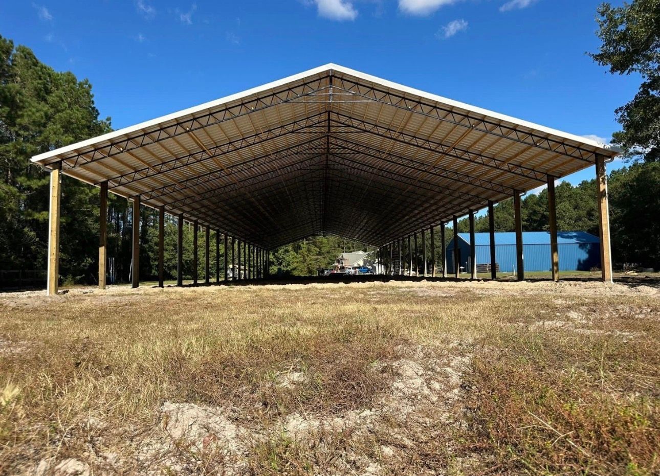 Large open-sided wooden structure with a white metal roof in a field. Blue sky and trees in the background.