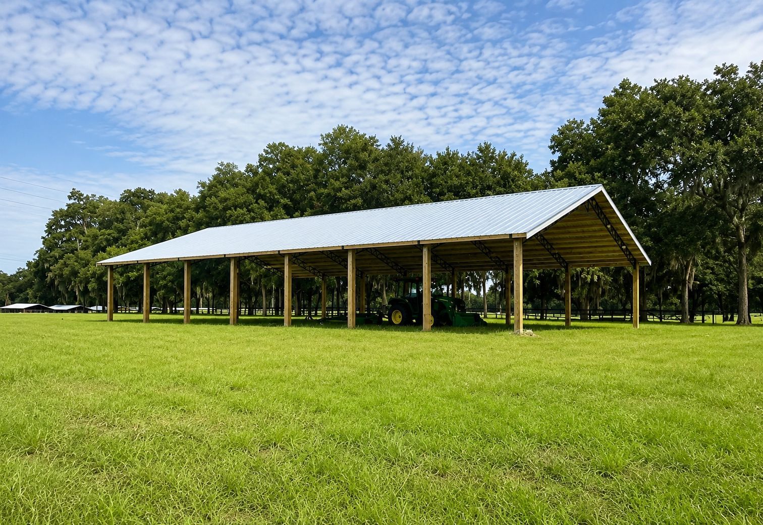 Large open-air shelter with a metal roof supported by wooden posts set in concrete, on a grassy field under a blue sky.