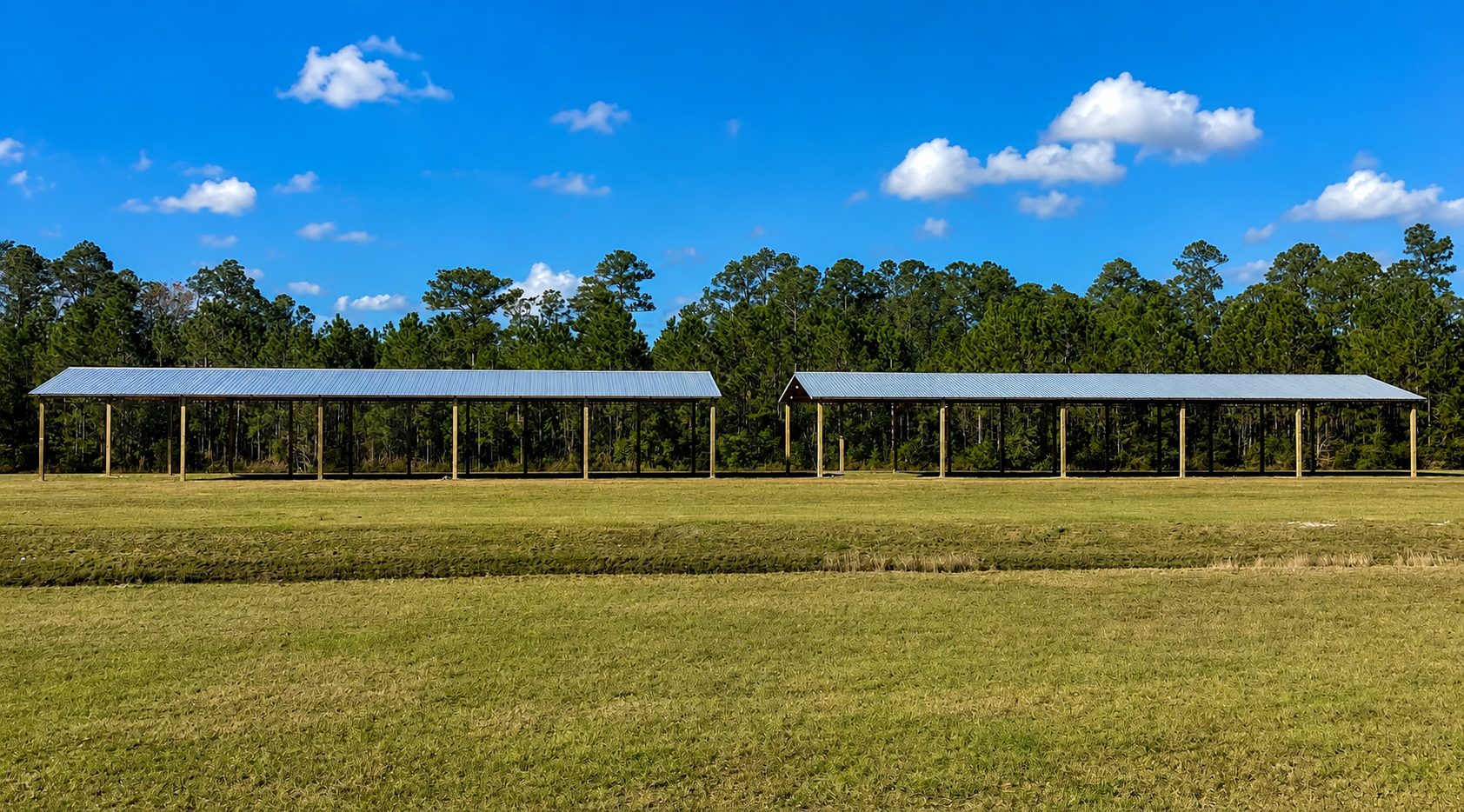 Two long, open-sided structures with metal roofs in a grassy field, trees in the background, blue sky.