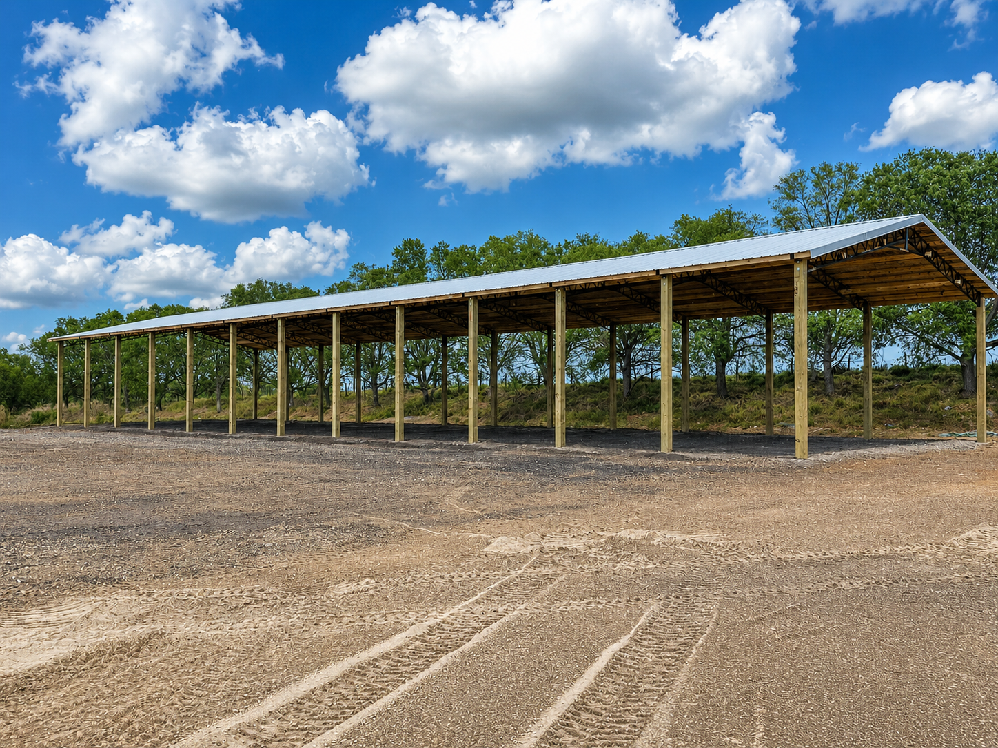 Two long, open-sided shelters with metal roofs in a field, trees in the background, blue sky.