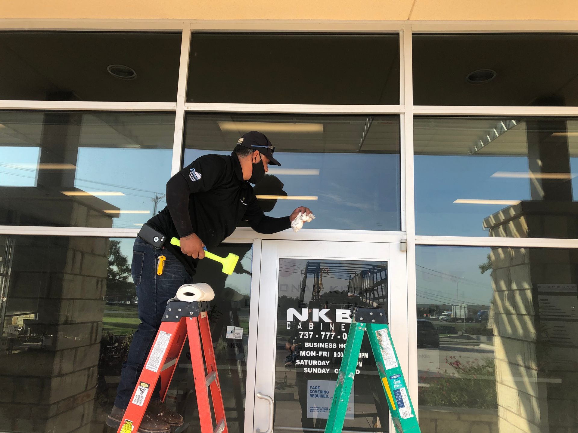 Man on a ladder removing window film from a building entrance. Wearing a mask, he holds a scraper.