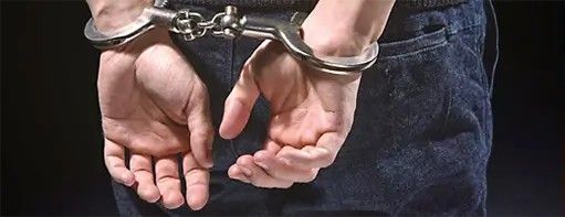 Person's hands cuffed behind their back, wearing jeans, against a dark background.