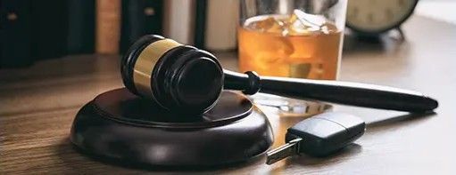 Gavel and car keys on a table next to a glass of whiskey, suggesting a DUI or legal issue.