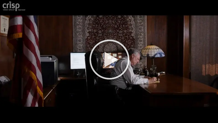 Man sitting at desk in an office with American flag, looking at paperwork.