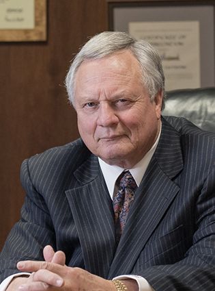 Man in suit, seated at desk, looking at the viewer with a stern expression.