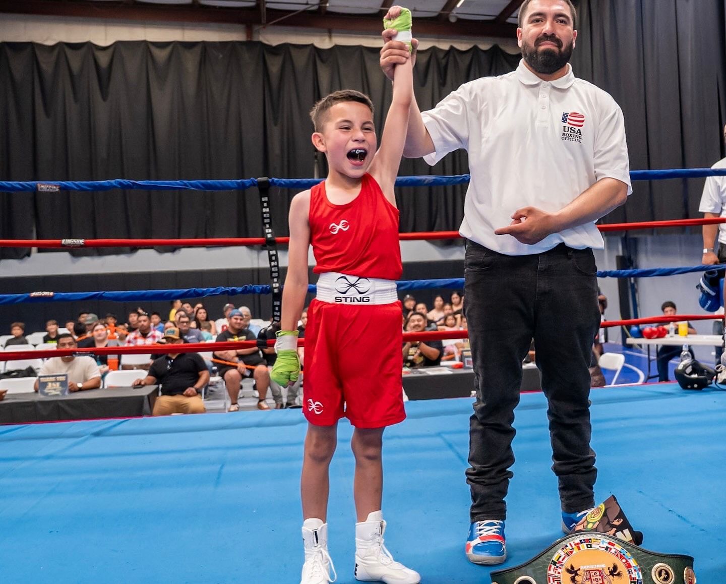 Young boxer in red outfit raises arm, celebrating victory. Referee points, championship belt on floor.