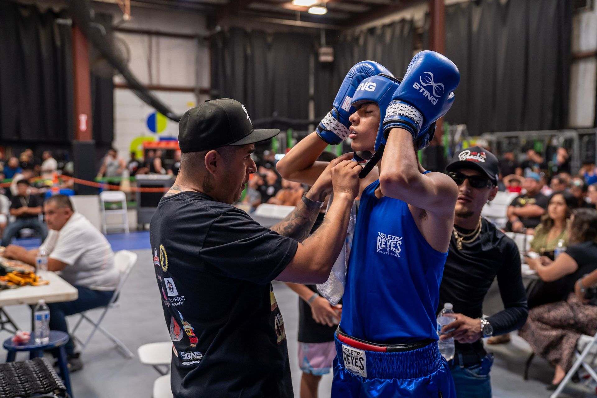 Boxer in blue getting headgear adjusted by a coach at a boxing match. Crowded indoor arena.