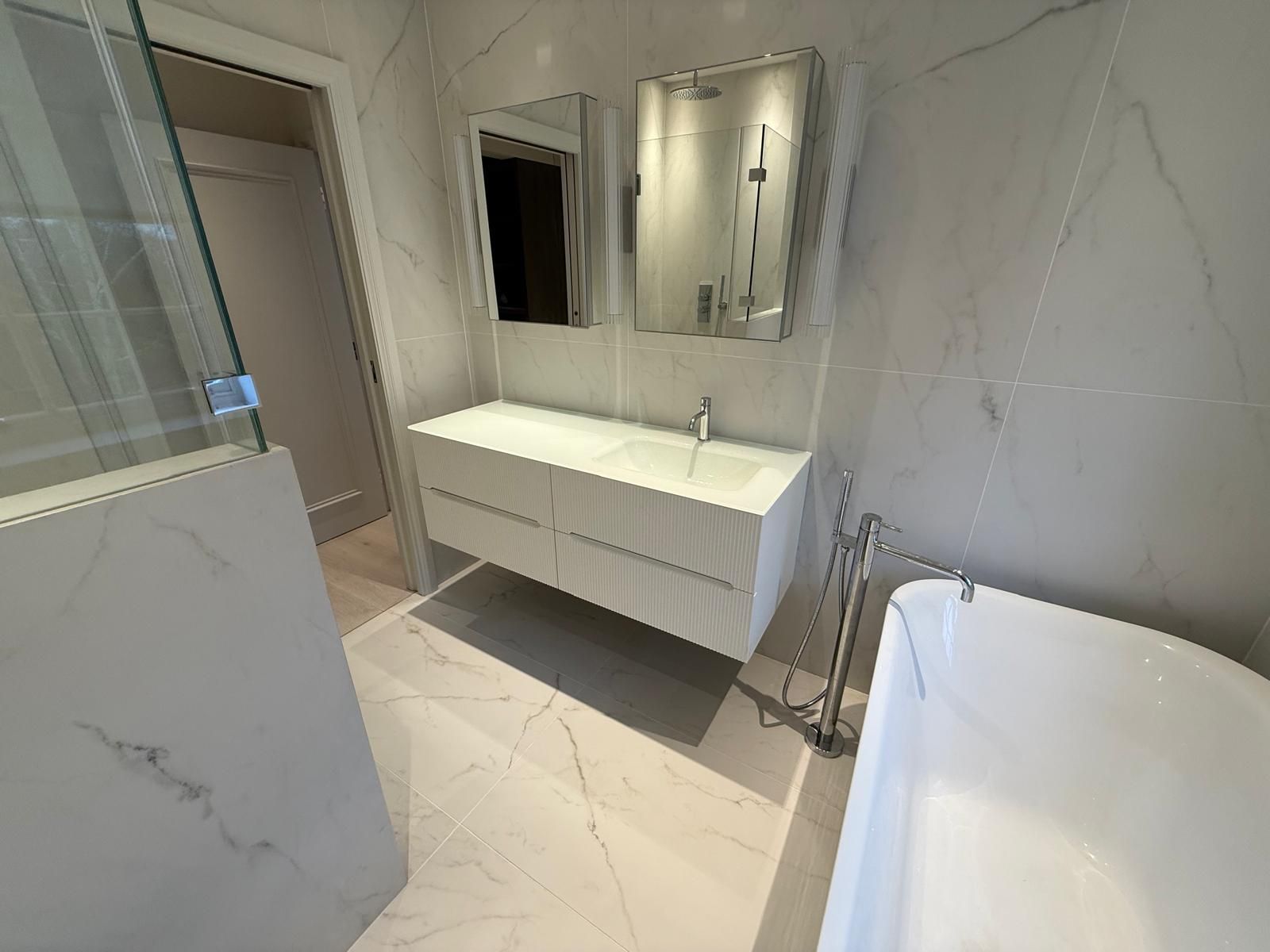 A modern bathroom featuring white marble walls, a floating double vanity with two mirrors, and a freestanding bathtub.