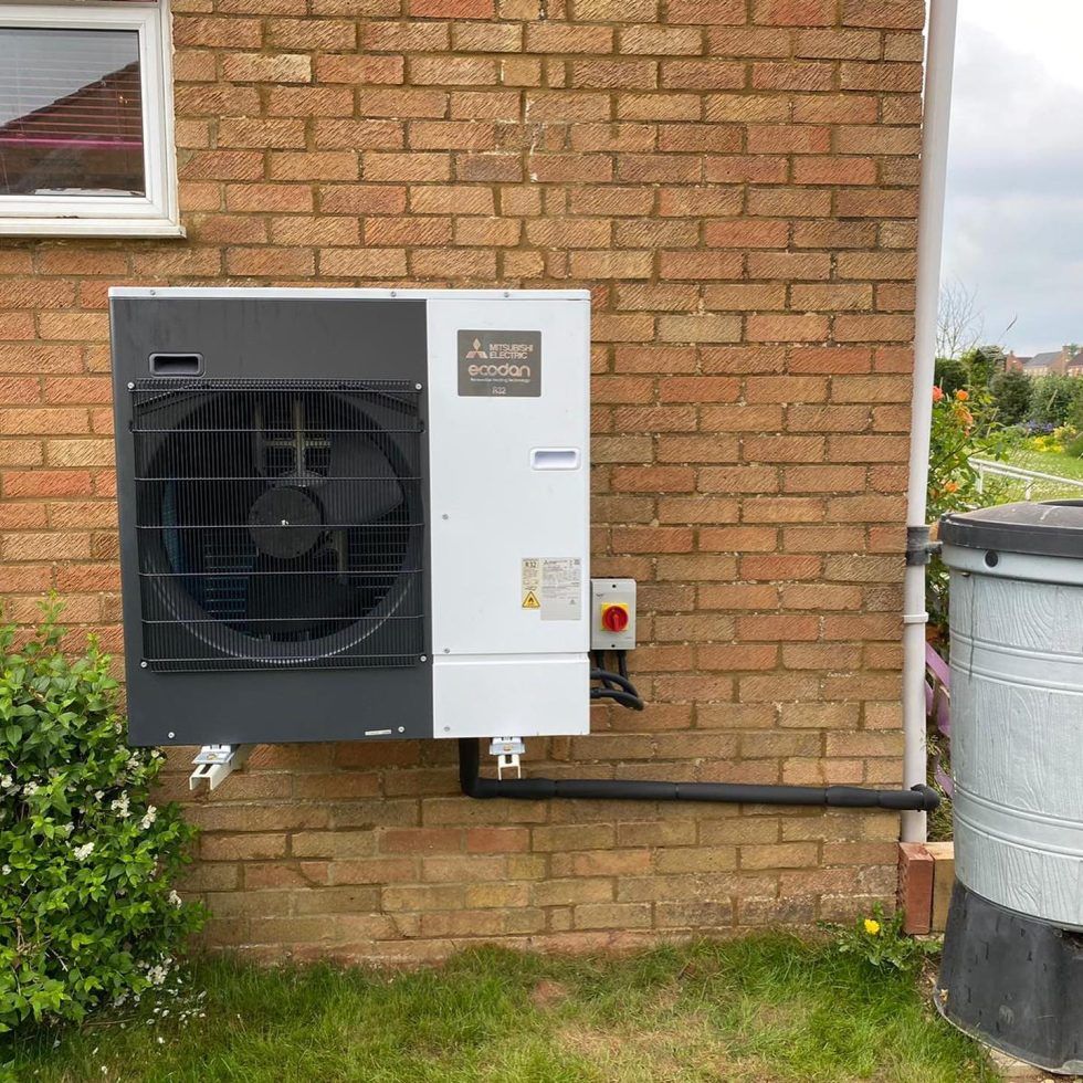 A Mitsubishi air source heat pump unit mounted on an exterior brick wall next to a vertical white pipe and metal bin.