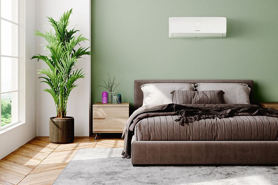 A modern bedroom with sage green walls, a brown bed, a large potted plant, and a white wall-mounted air conditioner.