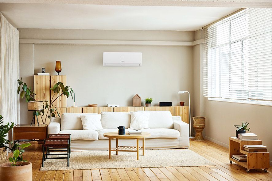 A bright, modern living room with a white sofa, a wooden coffee table, plants, and an air conditioning unit on the wall.